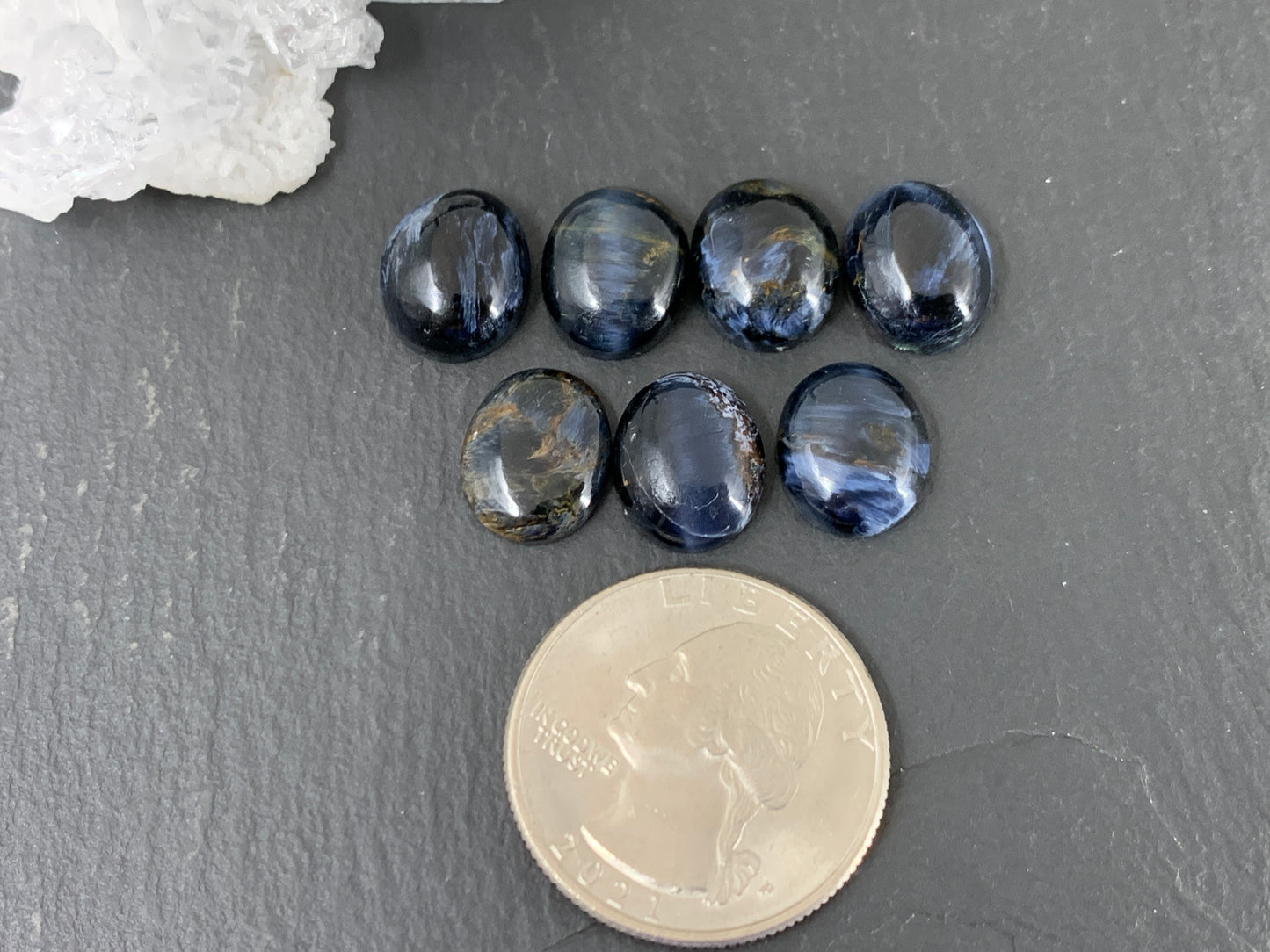 a collection of nine blue gemstones arranged on a gray surface, with a silver coin placed next to them.